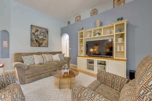A living room with a beige sofa, patterned cushions, a coffee table, and a TV on a white entertainment unit.