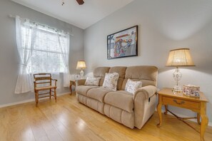 A living room with a beige sofa, two lamps, a wooden chair, a painting on the wall, and a window with sheer curtains.