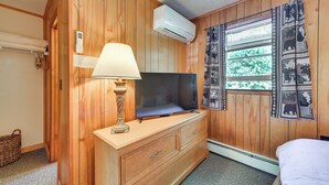 A room with wooden paneling, a television on a dresser, a lamp, a window with patterned curtains, and a wall-mounted air conditioner. - Gather Lakeside at Willsboro Inn on Lake Champlain (Willsboro)