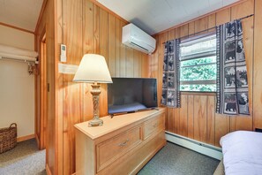 A room with wooden paneling, a television on a dresser, a lamp, a window with patterned curtains, and a wall-mounted air conditioner. - Gather Lakeside at Willsboro Inn on Lake Champlain (Willsboro)