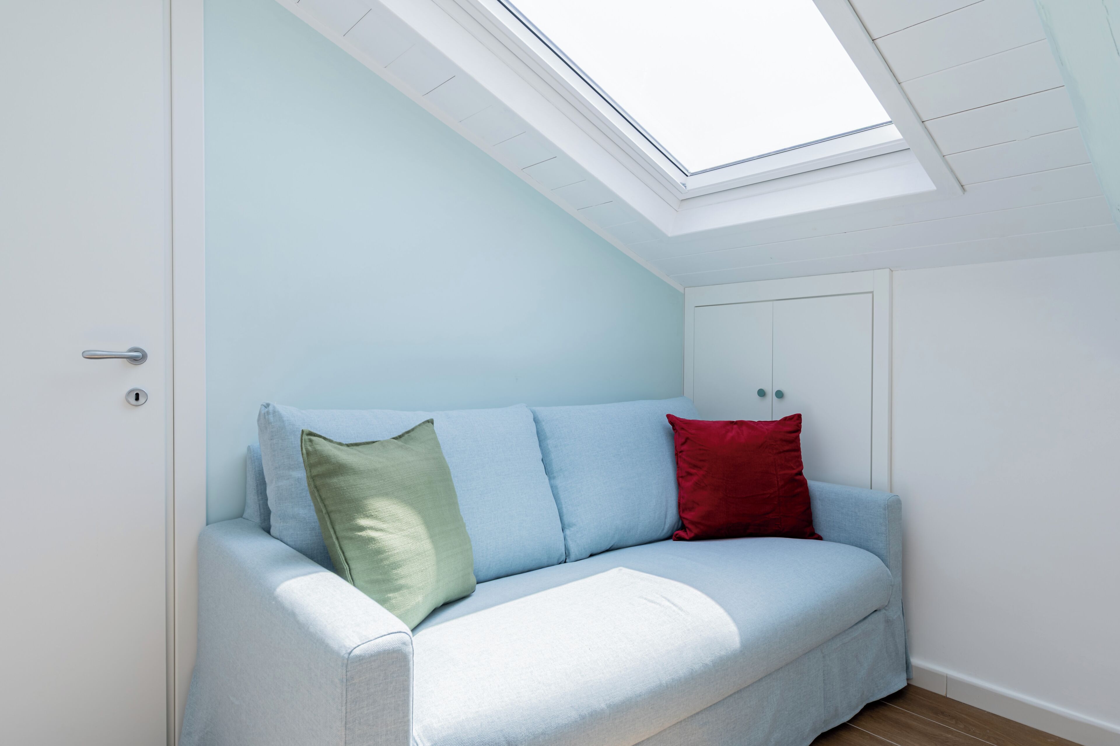 A light blue sofa with two pillows, one green and one red, in a room with a skylight.