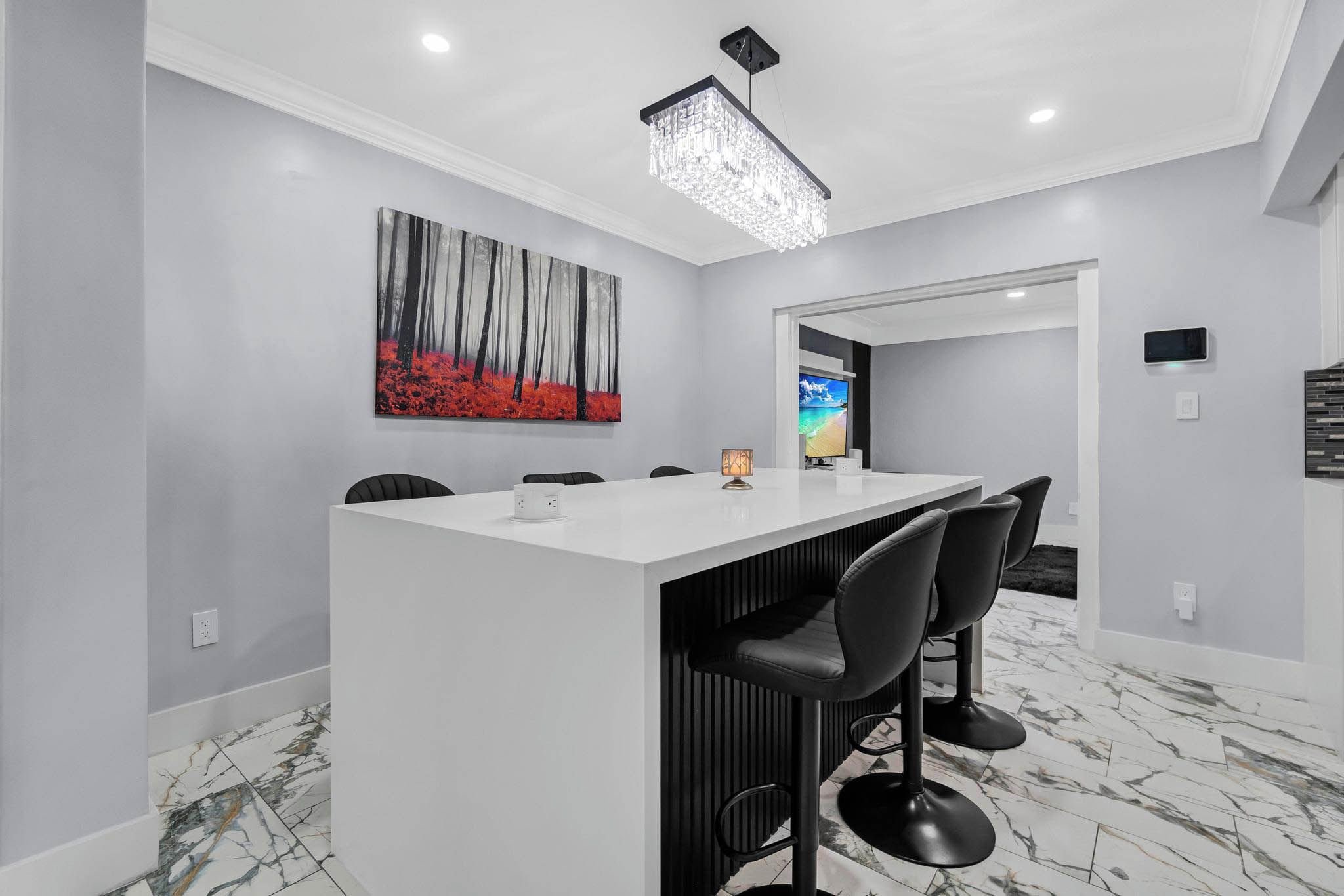 A modern kitchen with a white countertop, black bar stools, a chandelier, and a painting on the wall.