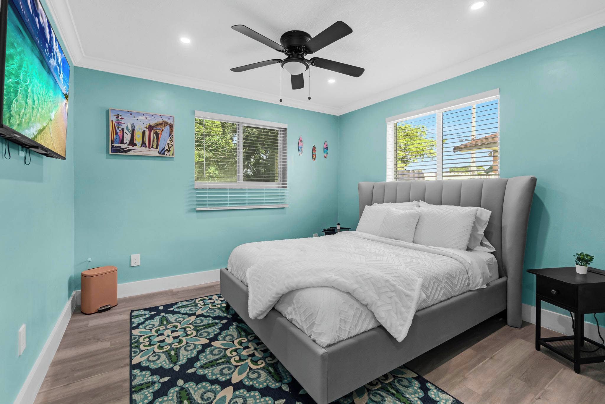 A bedroom with a bed, a ceiling fan, a window with blinds, and a wall-mounted TV.