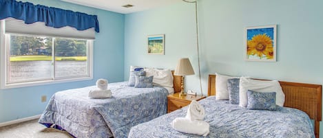 A bedroom with two beds, a ceiling fan, a window with a view of a body of water, and a painting of sunflowers.