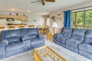 Ein Wohnzimmer mit zwei blauen Sofas, eine Essecke mit Tisch und Stühlen, eine Küche mit Holzschränken und ein großes Fenster mit Vorhängen.