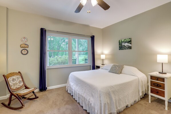 A bedroom with a large bed, a rocking chair, a window with curtains, a ceiling fan, and a nightstand with a lamp.