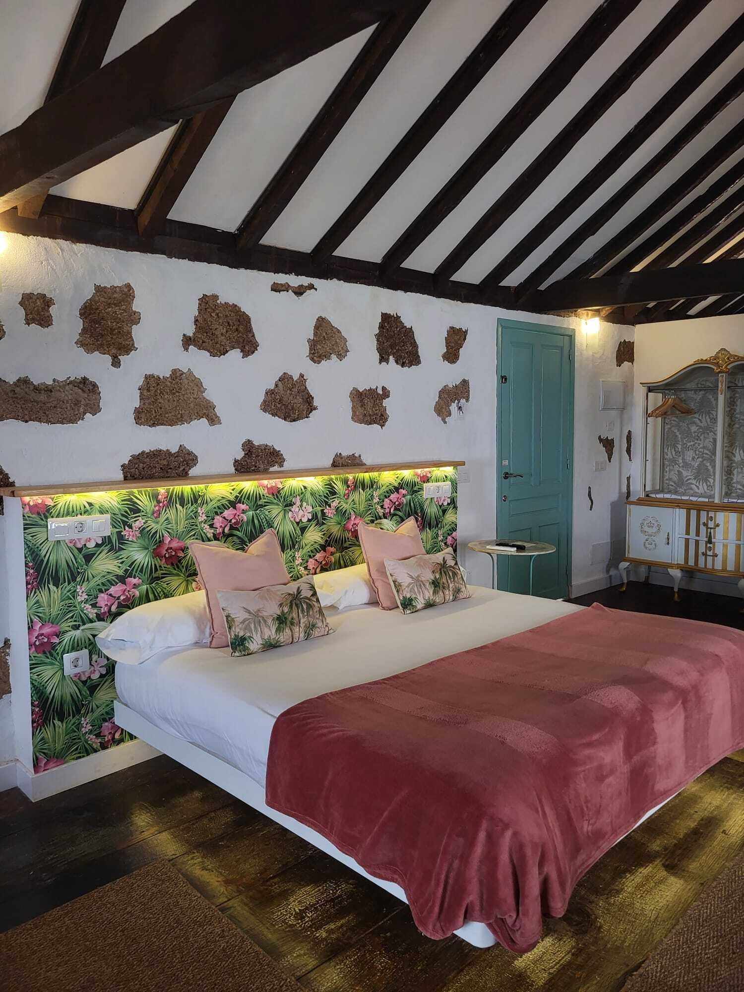 Un dormitorio con cama, cabecera de papel tapiz floral, puerta color esmeralda y mesita de noche pequeña.