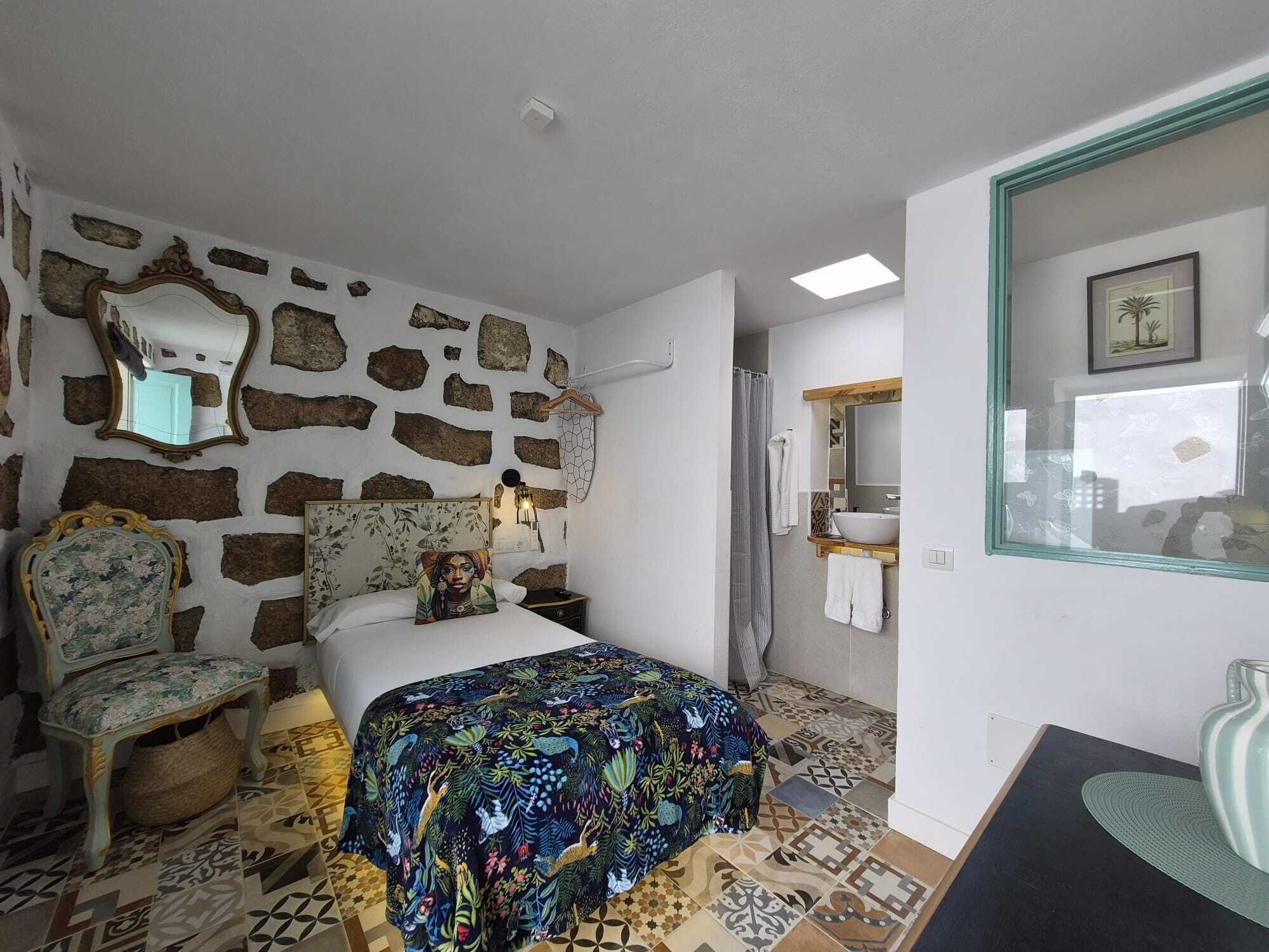 A room with a stone wall, a bed with a colorful bedspread, a chair with floral upholstery, a bedside table, and a mirror.
