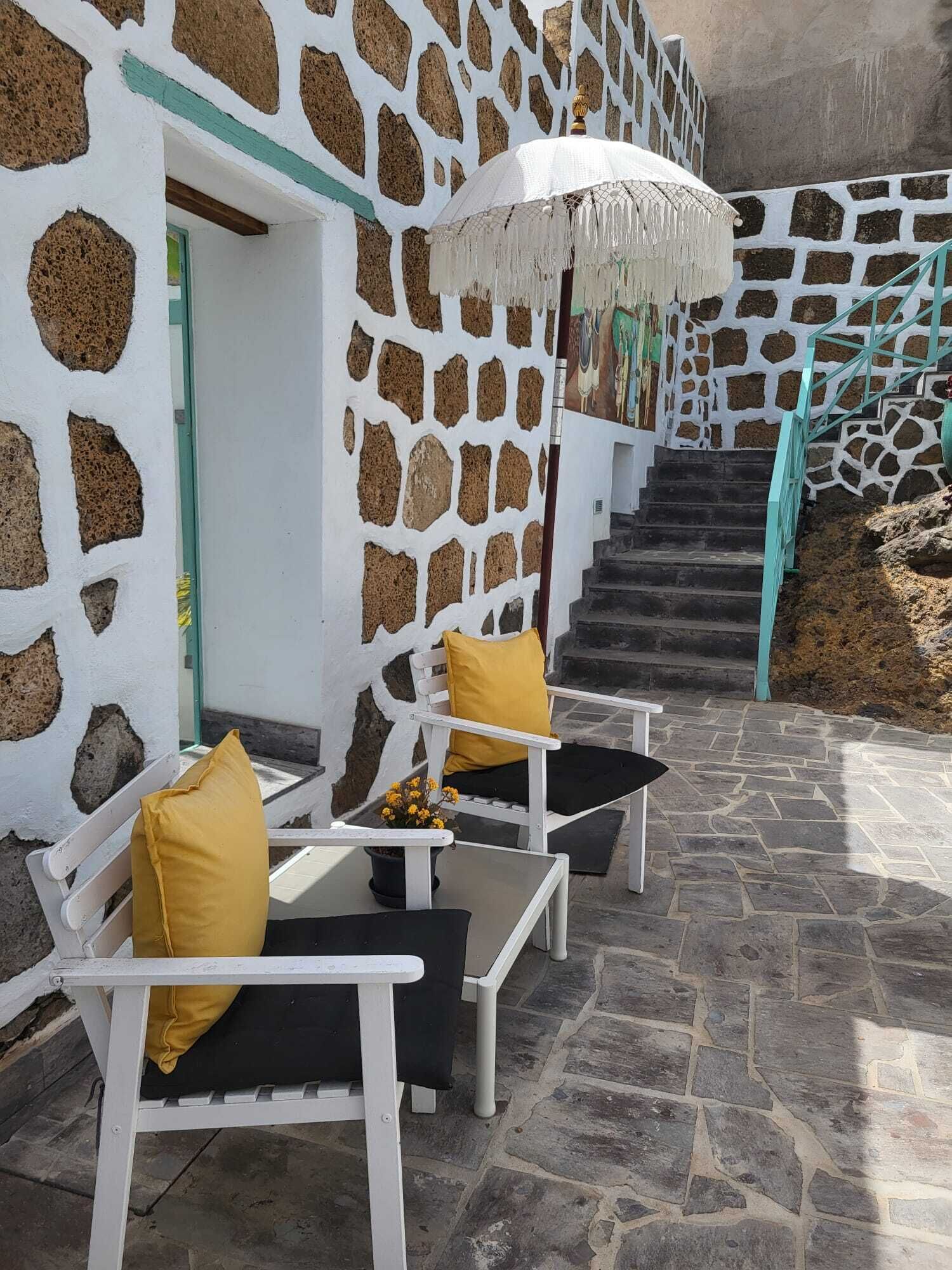 Un bâtiment aux murs de pierre, avec un escalier menant à une porte. Il y a des espaces extérieurs aménagés avec des chaises et des coussins.