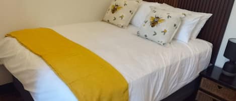 A bed with white bedding and a yellow throw blanket, two pillows with a bee design, a brown headboard, a nightstand with a lamp, and a woven rug.