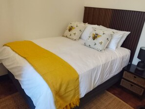 A bed with white bedding and a yellow throw blanket, two pillows with a bee design, a brown headboard, a nightstand with a lamp, and a woven rug.