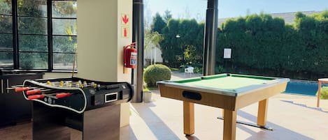 A pool table and foosball table in a room with large windows and a view of a swimming pool and greenery.