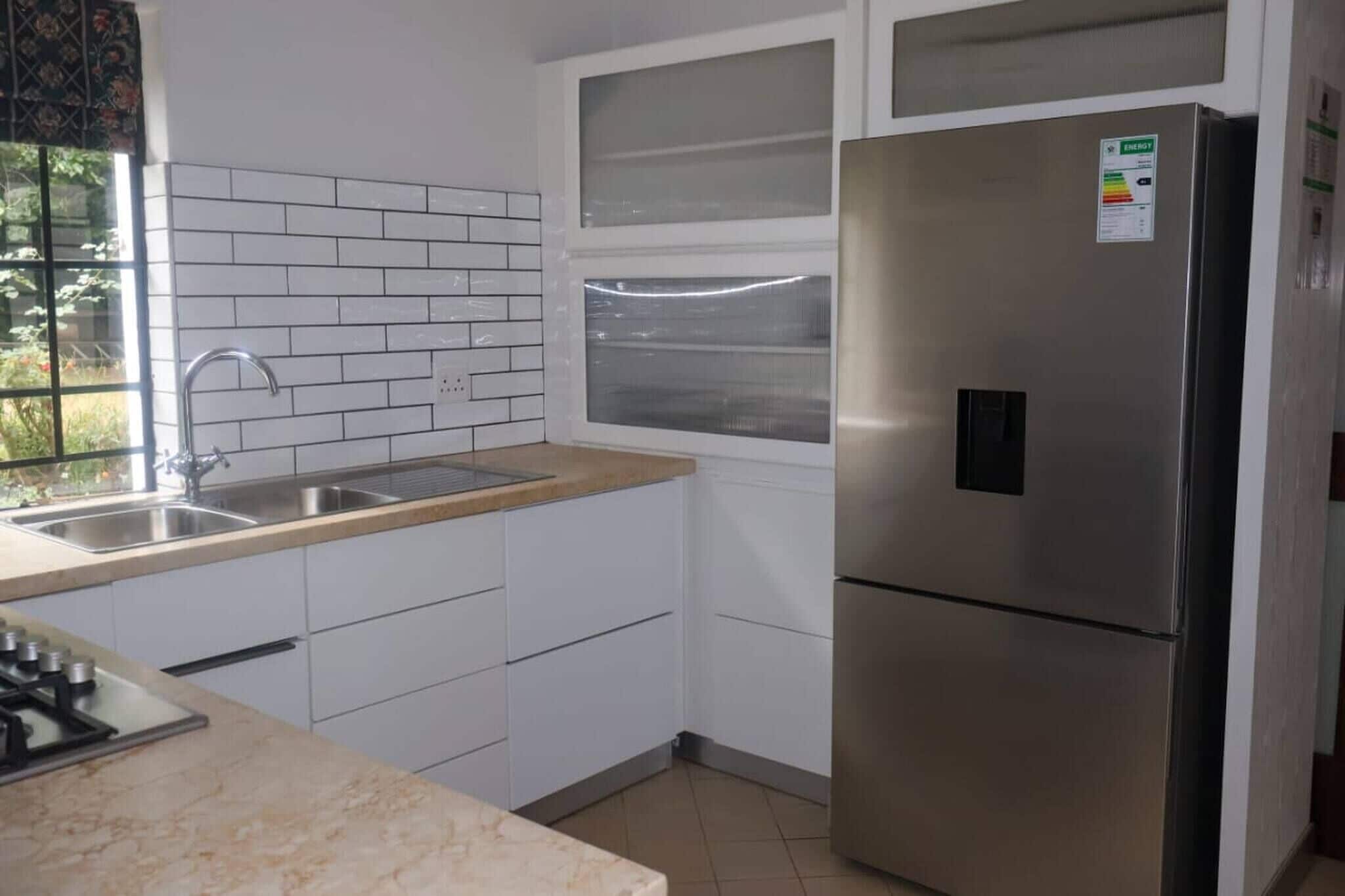 Una cocina con armarios blancos, un refrigerador de acero inoxidable, una ventana y un lavamanos.