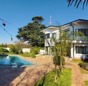 A two-story house with a swimming pool, surrounded by greenery and a clear sky.