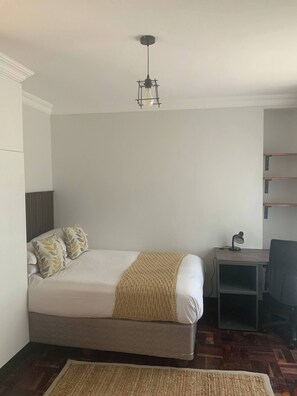 Una cama individual con cabecera, una mesita de noche con lámpara, una silla y un tapete tejido sobre piso de madera.