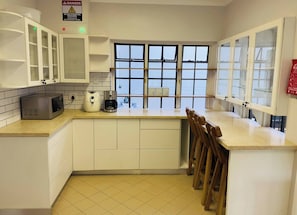 Una cocina con armarios blancos, microondas, cafetera y ventana con persianas.