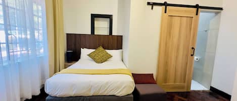 A hotel room with a bed, a bedside table, a mirror, a door, and a view of the outside.