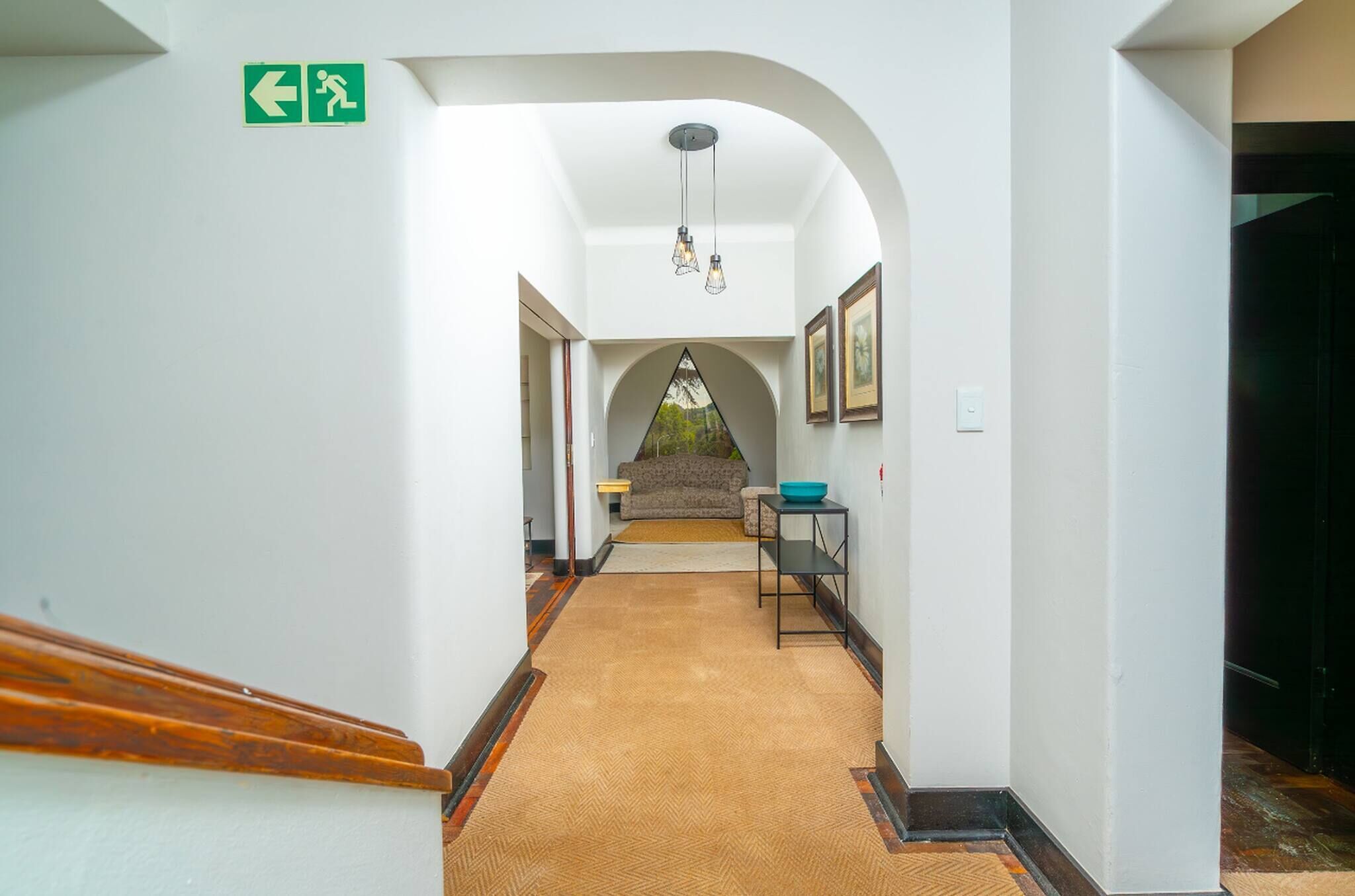 A hallway with a carpeted floor, a staircase, a sofa, a table with a bowl, framed pictures on the wall, and an exit sign.