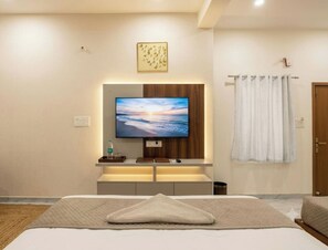 Habitación de hotel moderna con una cama, un televisor de pantalla plana montado en un panel de madera, una mesita de noche con una botella, vasos y un control remoto, y una cortina blanca en la ventana.