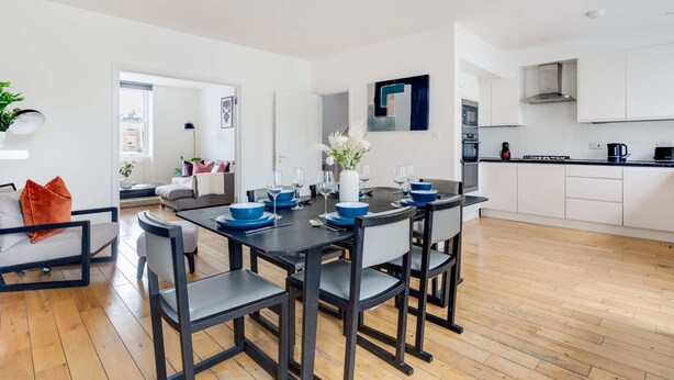 Een moderne keuken met witte kasten, een zwart aanrecht en een eethoek met een zwarte tafel en stoelen.