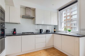 A modern kitchen with white cabinets, a gas stove, a coffee maker, and a window with a view of buildings. - Cozy 3BR Flat By Hyde Park (London)
