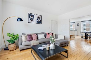 Ein modernes Wohnzimmer mit einem grauen Sofa, einem schwarzen Couchtisch und einer Stehlampe. Durch eine offene Tür ist ein Essbereich mit Tisch und Stühlen zu sehen.