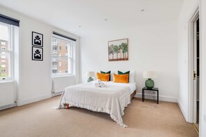 A bedroom with a bed, a nightstand, a lamp, a radiator, a window, and a painting on the wall. - Cozy 3BR Flat By Hyde Park (London)
