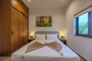 Habitación de hotel con una cama grande, un armario, un cuadro de un bosque, dos mesitas de noche con lámparas y una ventana con vistas a hojas de palmera.