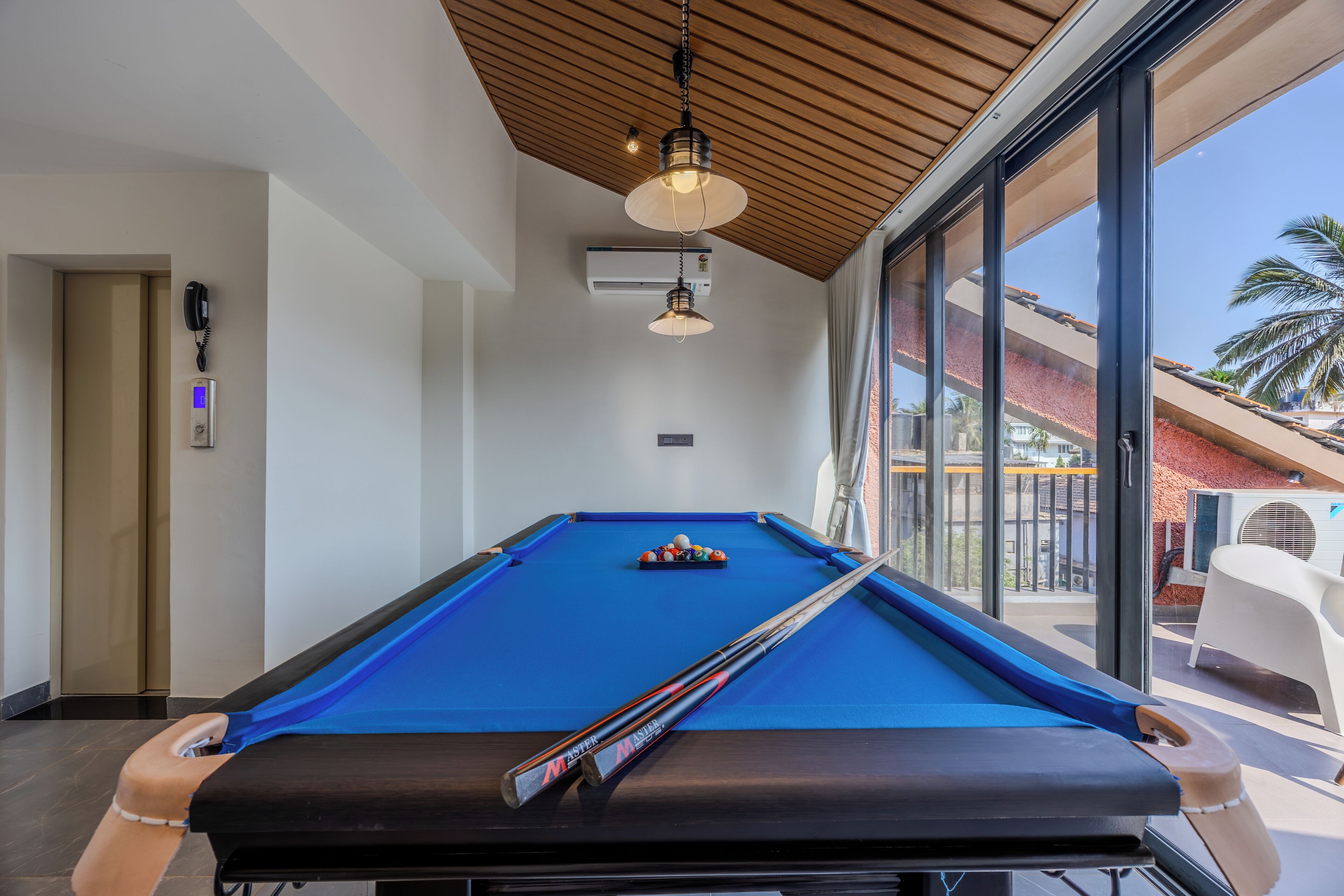 A pool table with pool cues and balls, a telephone, and an air conditioning unit in a room with large windows.