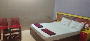 A hotel room with a bed, two red chairs, a small table, and a nightstand with a lamp.