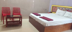 A hotel room with a bed, two red chairs, a small table, and a nightstand with a lamp.