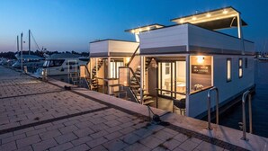Une propriété située au bord de l’eau, dotée d’un design moderne, comprenant un espace extérieur couvert, un escalier menant à l’entrée et une enseigne indiquant qu’il s’agit d’un pavillon en bord de mer.