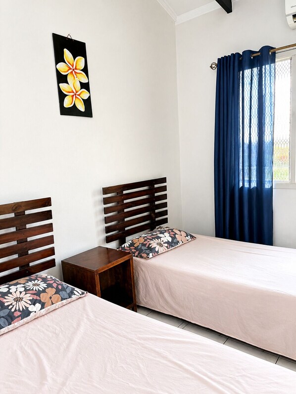 A room with two beds, a small wooden nightstand, a wall-mounted picture, and a window with blue curtains.