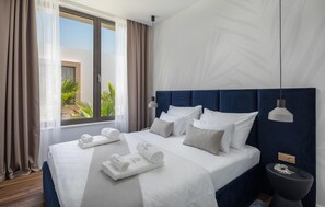 A hotel room with a large bed, a bedside table, a nightstand, a window with curtains, and a view of greenery outside. - Luxurious home in Tkon (Tkon)