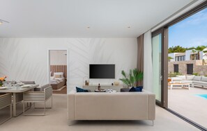 Ein modernes Wohnzimmer mit einer Couch, einem Essbereich, einem an der Wand montierten Fernseher und Blick auf den Außenpool.