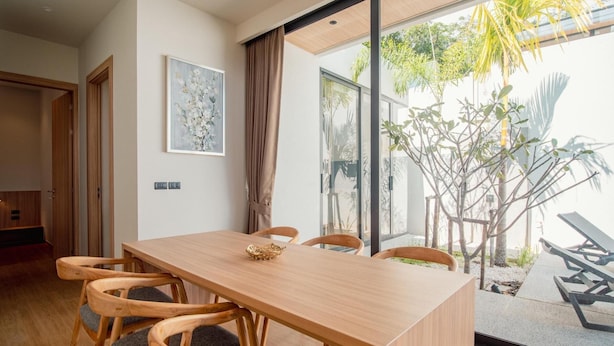 A modern dining area with a wooden table and chairs, a painting on the wall, and a view of the outdoors. - Villa Kauri 3BR at Aileen Naithon (Sa Khu)