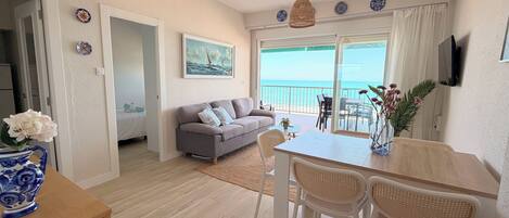 A living room with a sofa, a dining table with chairs, a TV mounted on the wall, and a view of the sea through the glass door.