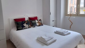 Ett hotellrum med en stor säng, vita sängkläder, två röda kuddar, ett fönster med utsikt över byggnader och en vit garderob.