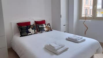 A hotel room with a large bed, white bedding, two red pillows, a window with a view of buildings, and a white wardrobe.