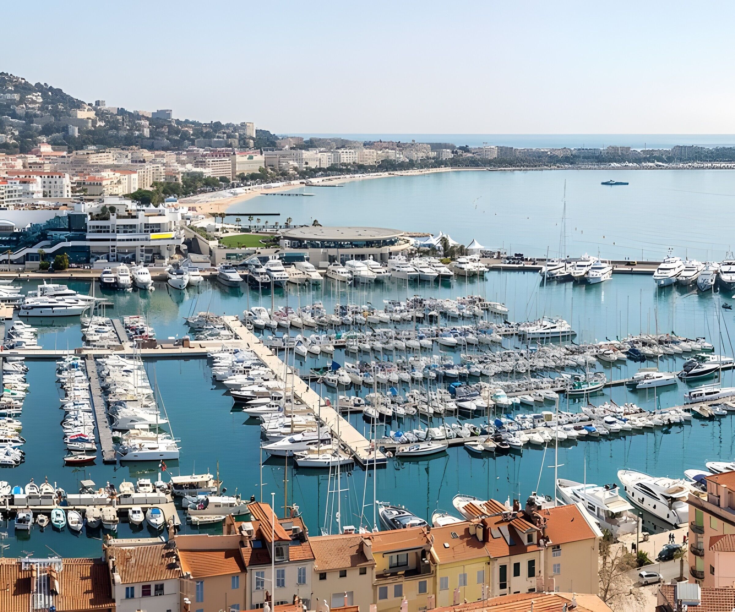 A marina with numerous boats docked, surrounded by buildings and a beach in the background.