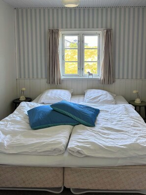 A bed with white linens and two blue pillows, a window with curtains, and a striped wall. - Summer house with wilderness bath near Henne (Oksbøl)