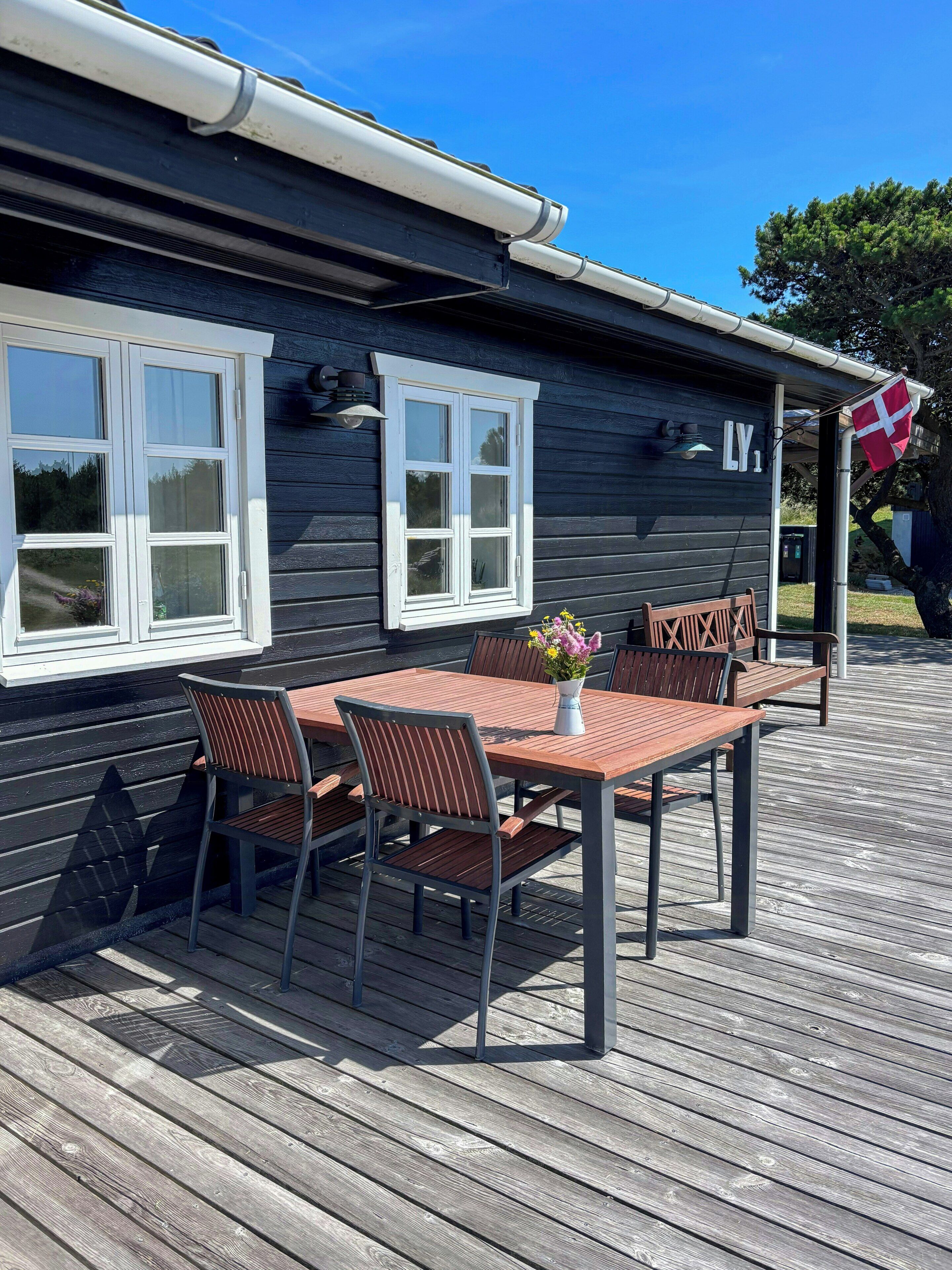 Une terrasse en bois avec une table et des chaises, un drapeau et un bâtiment doté de fenêtres et d’une enseigne.