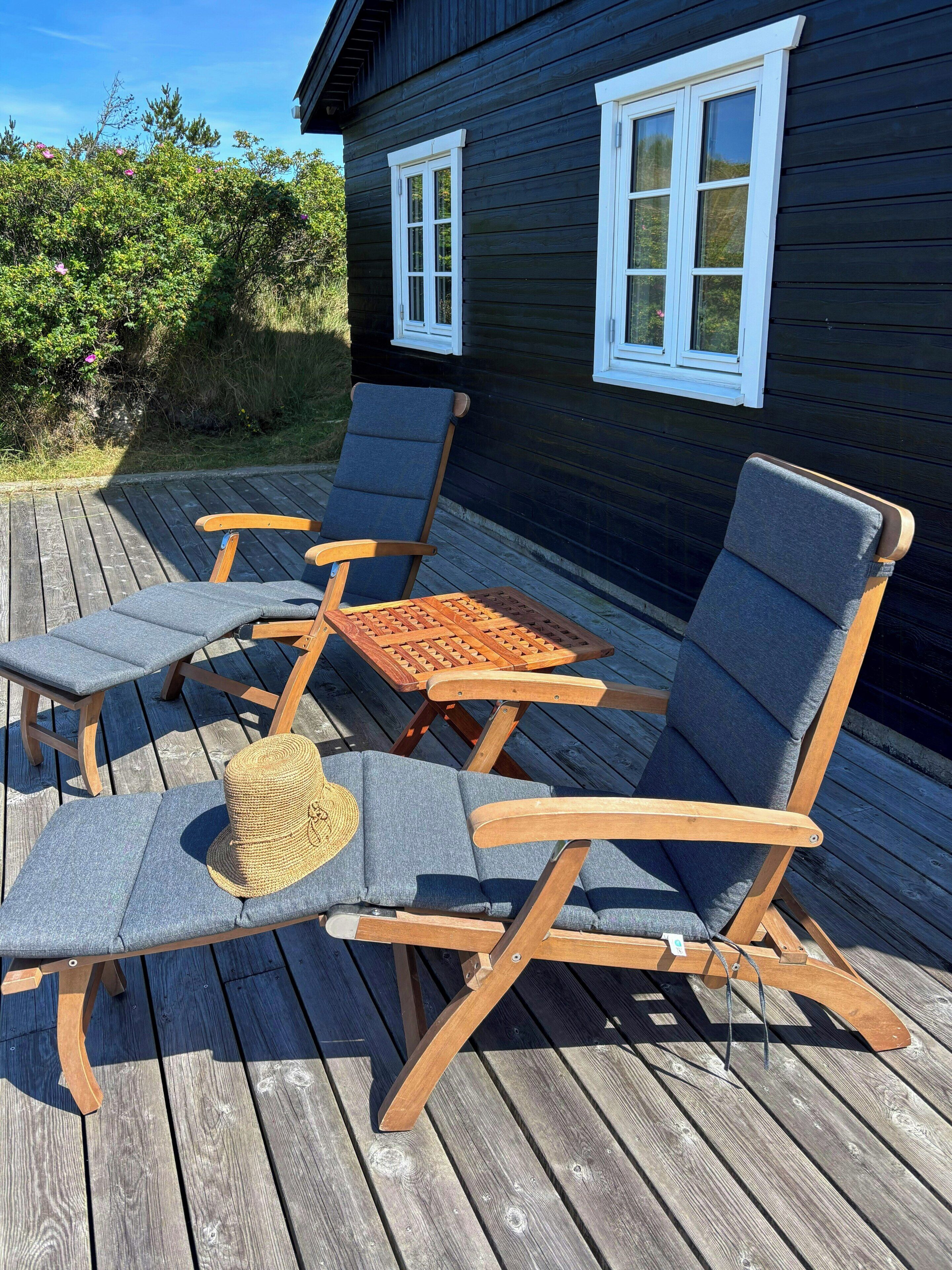 A deck with wooden furniture including lounge chairs and a table, adjacent to a dark wooden building with white-framed windows.