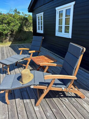 A deck with wooden furniture including lounge chairs and a table, adjacent to a dark wooden building with white-framed windows. - Summer house with wilderness bath near Henne (Oksbøl)
