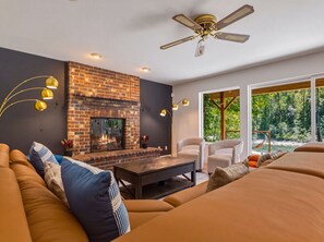 A modern living room with a brick fireplace, a ceiling fan, and a large window offering a view of a forest. - Riverfront Resort w/ Spa~Firepit~Hammock~Theater~Games~Sleeps 20~Amazing Views (Gold Bar)