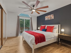 Ein Schlafzimmer mit Bett, Deckenventilator, Nachttisch mit Lampe und Blick ins Freie durch ein großes Fenster.