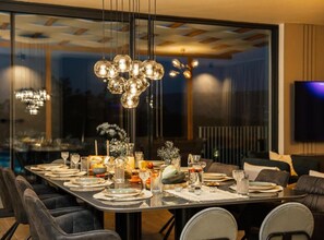 Ein moderner Essbereich mit einem langen Tisch, der für eine Mahlzeit gedeckt ist, einer Chandelüster und einem an der Wand befestigten Fernseher.