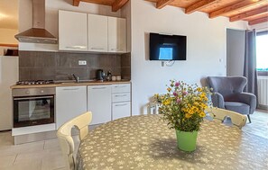 A compact kitchen with a stove, oven, and white cabinets. There's a dining table with a floral tablecloth and a green vase with flowers. A flat-screen TV is mounted on the wall above the table. - Stunning home in Pocekaji (Pocekaji)