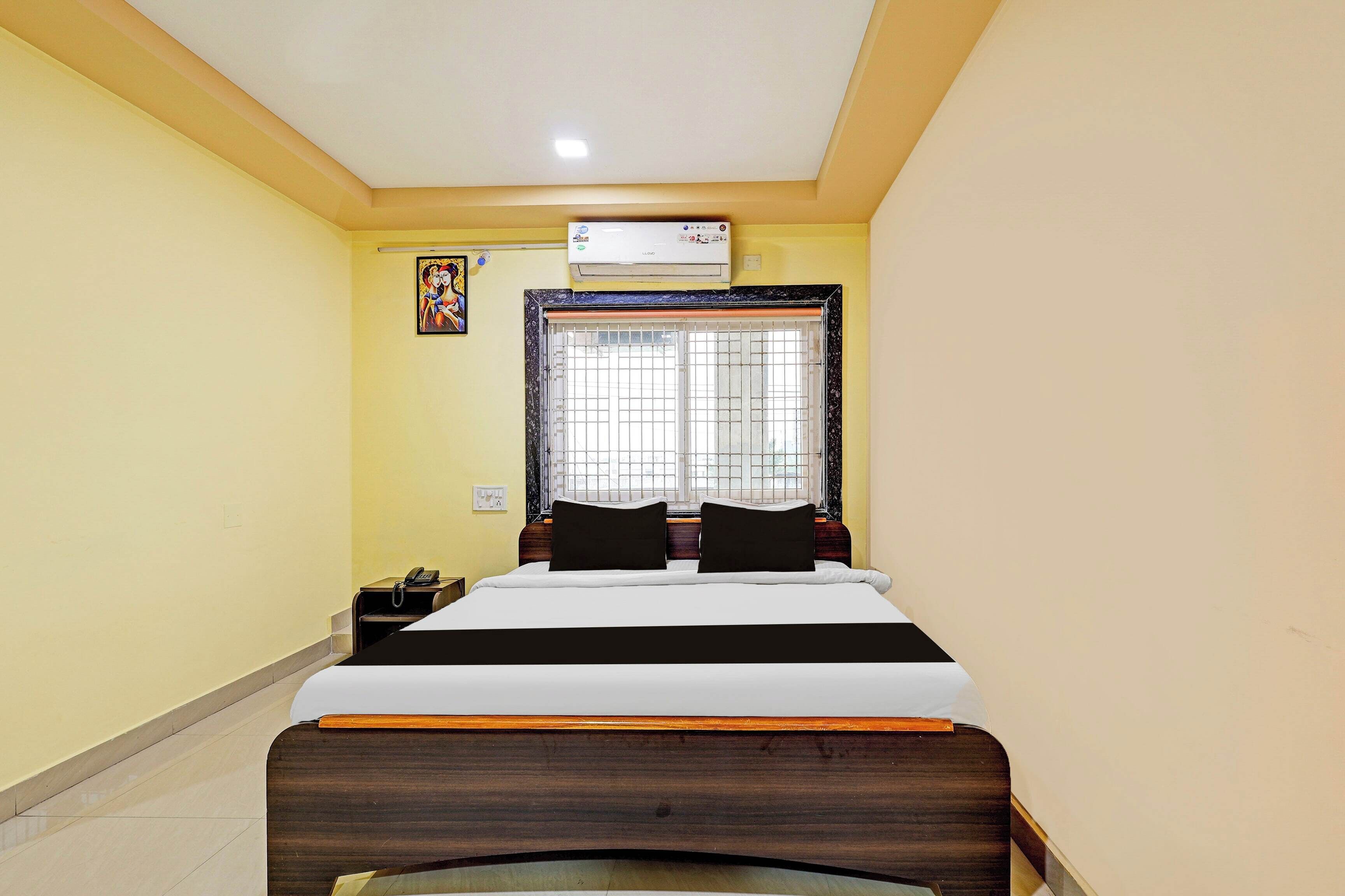 A hotel room with a bed, a window with bars, an air conditioning unit, and a telephone on a side table.