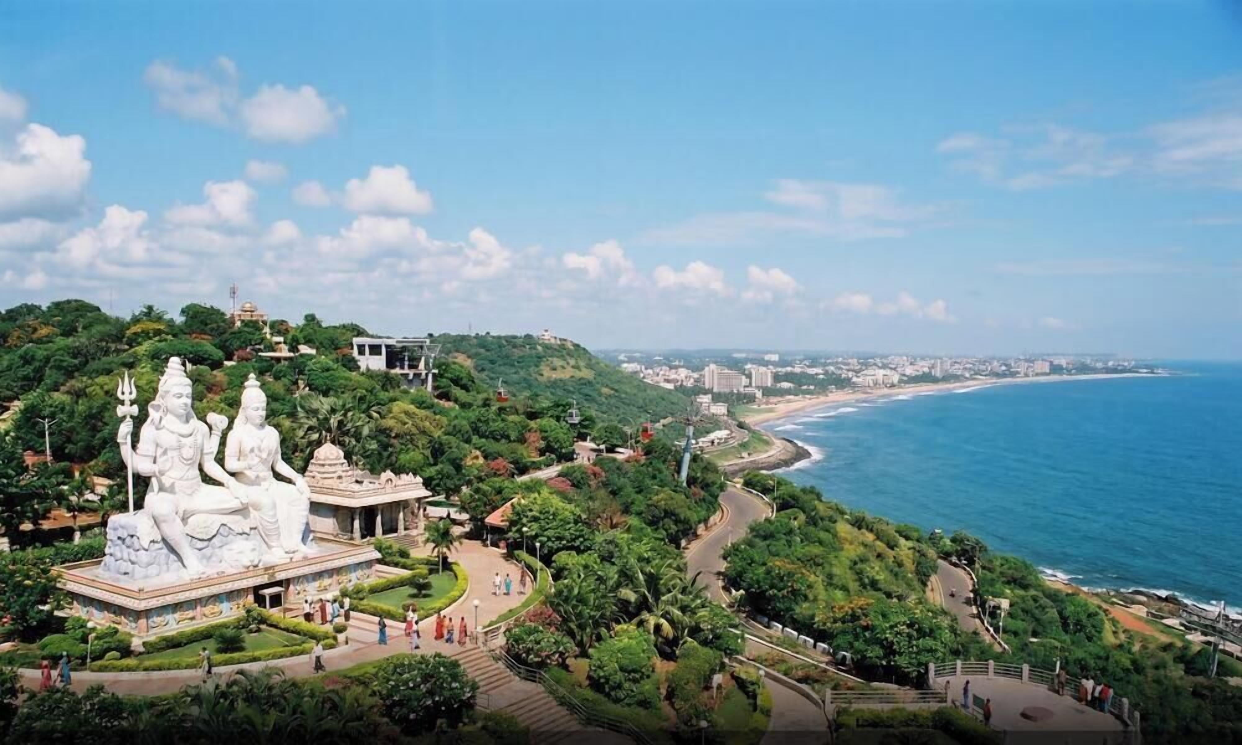 A coastal landscape with a temple featuring large white statues, a winding road, and a beach.